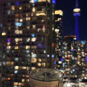 K&ouml;nnte beinhalten: Duftkerze in einem transparenten Glas, mit einem hellblauen Blumendesign. Das Etikett tr&auml;gt die Aufschrift "Scented Candle". Im Hintergrund eine n&auml;chtliche Stadtlandschaft mit beleuchteten Geb&auml;uden und einem hohen Turm.
