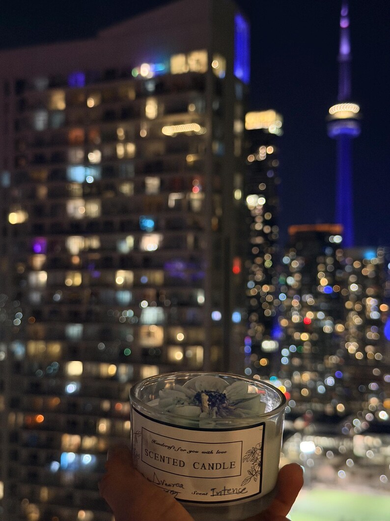 K&ouml;nnte beinhalten: Duftkerze in einem transparenten Glas, mit einem hellblauen Blumendesign. Das Etikett tr&auml;gt die Aufschrift "Scented Candle". Im Hintergrund eine n&auml;chtliche Stadtlandschaft mit beleuchteten Geb&auml;uden und einem hohen Turm.