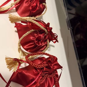K&ouml;nnte beinhalten: Drei rote Satin-Kordelzugbeutel mit goldenen Seilgriffen und Quasten. Die Beutel sind mit roten B&auml;ndern gebunden und haben eine gl&auml;nzende, reflektierende Oberfl&auml;che. Der Hintergrund ist eine wei&szlig;e Oberfl&auml;che.