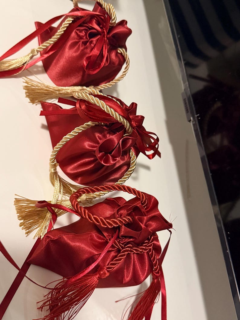 K&ouml;nnte beinhalten: Drei rote Satin-Kordelzugbeutel mit goldenen Seilgriffen und Quasten. Die Beutel sind mit roten B&auml;ndern gebunden und haben eine gl&auml;nzende, reflektierende Oberfl&auml;che. Der Hintergrund ist eine wei&szlig;e Oberfl&auml;che.