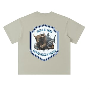May include: A light beige short-sleeve t-shirt featuring a graphic on the back. The design shows a yak and a shark hybrid creature surrounded by beer bottles and cans, enclosed in a shield shape with the text YAK & ATTACK NEVER MISS A ROUND.