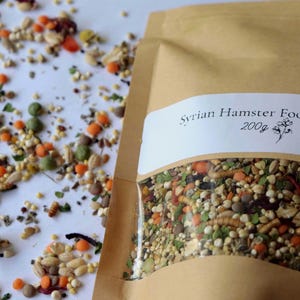 May include: A brown paper bag of Syrian hamster food mix, with a clear window showing the contents. The mix includes various seeds, grains, and dried insects. The label reads "Syrian Hamster Food Mix 200g". Some food is scattered around the bag.