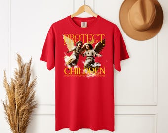 May include: A red short-sleeve t-shirt featuring a graphic print of two winged figures with the yellow text "PROTECT CHILDREN" above and below the image. The shirt has a crew neckline and is displayed on a wooden hanger.