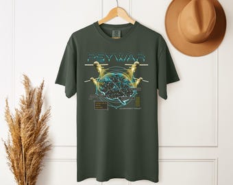 May include: A dark olive green short-sleeve t-shirt featuring a graphic print with the text PSYWAR. The design depicts a human brain surrounded by four helicopters with spotlights, technical lines, and text including GOVERNMENT TARGET.