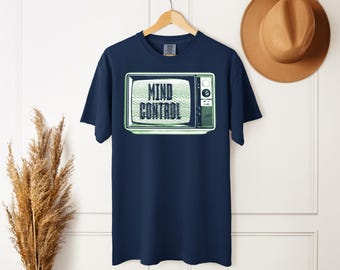 May include: A navy blue short-sleeve t-shirt featuring a graphic print of a vintage television set. The screen displays static noise and the text "MIND CONTROL" in a bold, dark font. The television is light green with a side control panel.