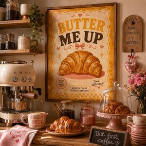 Puede incluir: Una acogedora escena de cocina con una máquina de espresso de estilo vintage, un póster enmarcado "Butter Me Up" con un cruasán y cruasanes frescos en un soporte rosa para pasteles. La escena incluye tazas de café, una toalla rosa y una pequeña pizarra.