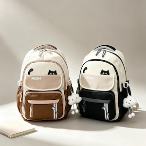 May include: Two backpacks in cream and brown or cream and black, featuring a cat graphic with the text MEOW, a front flap pocket, side pockets, and a white plush cloud charm attached to the side. Each bag has a top handle and zipper closures.