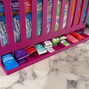 May include: A bright pink, 3D-printed organizer for drink mix packets and drink mix sticks. The organizer has multiple slots for different flavors, including Hawaiian Punch and Skittles. The bottom drawer holds individual packets.