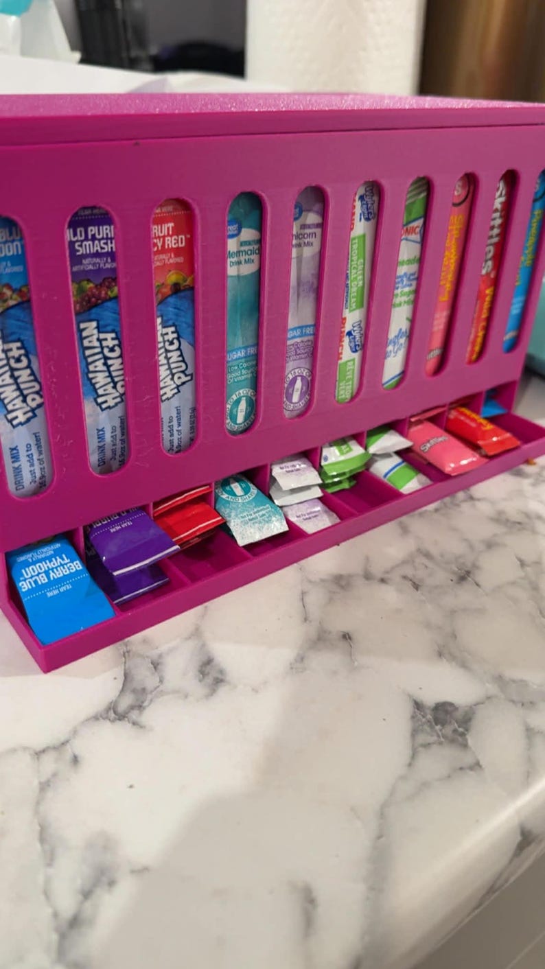 May include: A bright pink, 3D-printed organizer for drink mix packets and drink mix sticks. The organizer has multiple slots for different flavors, including Hawaiian Punch and Skittles. The bottom drawer holds individual packets.