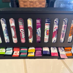 May include: A black dispenser filled with various flavored drink mix packets. The packets are in a row and include flavors like "Margaritaville", "Mermaid", "Crush", "Hawaiian Punch", "Unicorn", and "Sherbet".
