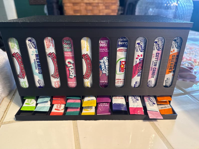 May include: A black dispenser filled with various flavored drink mix packets. The packets are in a row and include flavors like "Margaritaville", "Mermaid", "Crush", "Hawaiian Punch", "Unicorn", and "Sherbet".