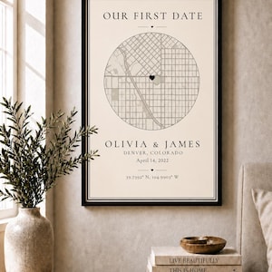 Puede incluir: Una impresión enmarcada con el texto "OUR FIRST DATE" sobre un mapa de Denver, Colorado. Los nombres "OLIVIA & JAMES" y la fecha "April 14, 2022" están debajo. La impresión se muestra en una mesa de madera con libros y un jarrón.
