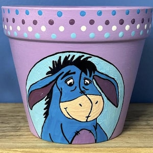 May include: A hand-painted, lavender-colored flower pot featuring a cartoon image of Eeyore from Winnie the Pooh. The pot has a blue and white polka dot design around the rim. The Eeyore image is set against a light blue background.
