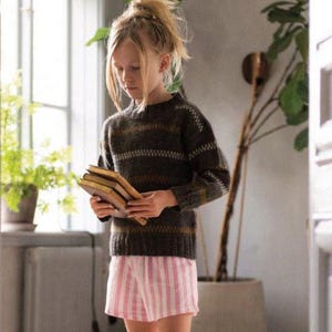 May include: A young person wearing a dark brown striped sweater and pink and white striped shorts. They are holding several books. The person is standing barefoot in a room with a window and a large potted plant.