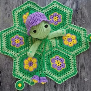 Puede incluir: Una manta de seguridad de ganchillo con una cabeza de tortuga verde claro y un gorro morado, unida a una base en forma de estrella hecha de cuadrados de abuela florales verdes, amarillos y morados. Incluye dos botones amarillos y flores moradas colgantes.