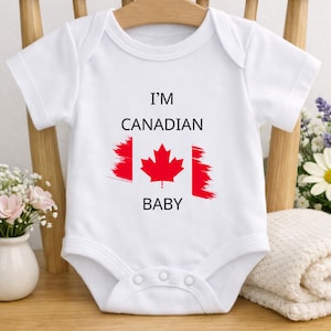 May include: A white short-sleeved baby bodysuit featuring the text I'M CANADIAN BABY in black lettering. Centered between the lines of text is a red maple leaf graphic with brushstroke-style red vertical bars on either side of the leaf.