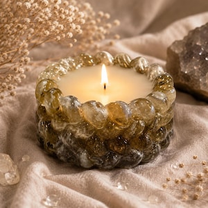 Pode incluir: Uma vela acesa num castiçal decorativo de vidro. A vela tem cera branca cremosa e uma chama trémula. O castiçal é feito de contas de vidro transparentes e âmbar. A vela está sobre um tecido bege.