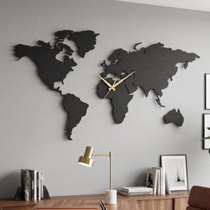 May include: A large black world map wall clock with gold-colored hands mounted on a gray wall. The map features a stylized silhouette of the continents, with the clock mechanism integrated into the central portion of the African continent.