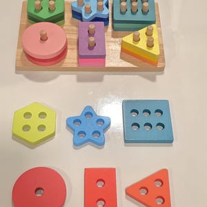 May include: A wooden shape sorter toy with a light brown base and colorful geometric shapes. The shapes include a hexagon, star, square, circle, rectangle, and triangle, each with corresponding pegs and holes. The colors are green, blue, pink, yellow, orange, and red.