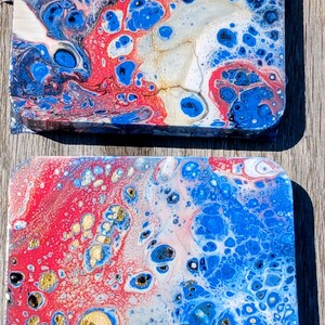 Puede incluir: Dos posavasos cuadrados con un diseño abstracto de vertido de acrílico fluido. La superficie muestra una mezcla vibrante de pintura roja, azul real, blanca y dorada metálica, creando patrones orgánicos de celdas y texturas marmoladas.