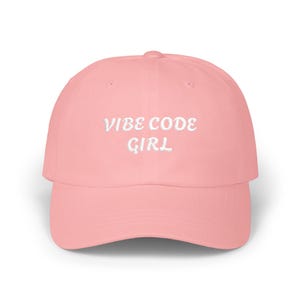 May include: A light pink baseball cap featuring the text VIBE CODE GIRL embroidered in white thread across the front panel. The cap has a structured crown, a curved brim, and a button at the top center, presenting a clean and simple design.
