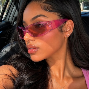 May include: Wraparound sunglasses featuring a translucent hot pink plastic frame and matching pink tinted lenses. The eyewear has a sleek, elongated shape that contours the face, with thick temples and a seamless bridge design in a vibrant magenta hue.