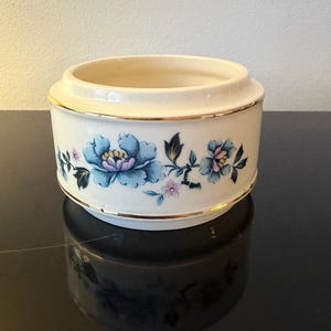 May include: A cream-coloured ceramic container with a floral design. The container features blue and pink flowers with gold trim around the top and bottom. The container is cylindrical with a wide opening.