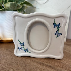 May include: A rectangular ceramic picture frame with a wavy edge and an oval centre opening. The frame is off-white and features painted green stems with small blue butterflies on the bottom left and right sides of the central oval opening.