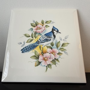 May include: A square white ceramic tile featuring a detailed illustration of a blue jay perched among light pink flowers and green leaves. The bird has blue and white plumage with black markings, and the floral arrangement is centred on the tile.