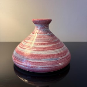 May include: A small, decorative vase with a bulbous body and a narrow neck. The vase has horizontal stripes of red, white, and light blue. The glossy finish reflects light, enhancing the colours and design. The vase is likely ceramic.