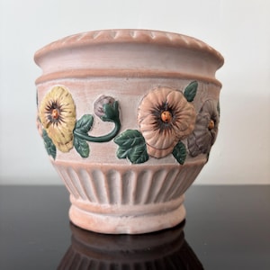 May include: A terracotta plant pot with embossed floral designs. The pot has a light pink base with vertical ribbed detailing and a scalloped rim. The flowers are in shades of yellow, peach, and purple with green leaves.