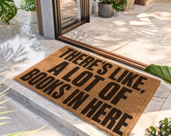 Puede incluir: Un felpudo rectangular de fibra de coco color canela con el texto en negro "THERE'S LIKE, A LOT OF BOOKS IN HERE". La alfombra presenta una textura de fibra natural y está colocada sobre un suelo de baldosas claras frente a una puerta de cristal.