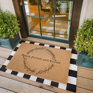May include: A rectangular doormat with the text "Speak Friend and Enter" in a circular design. The mat is made of a natural fiber material and is surrounded by a black and white striped border. Two green potted plants flank the door.