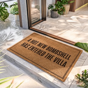 May include: A rectangular tan coir doormat with a black double-line border. The text A HOT NEW BOMBSHELL HAS ENTERED THE VILLA is printed in bold black capital letters across the center of the mat, positioned on a light-colored tiled entryway floor.