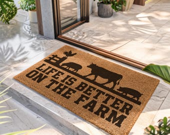 Puede incluir: Un felpudo rectangular de fibra de coco color canela con un diseño impreso en negro de un gallo sobre una valla, una oveja, una vaca y un cerdo. El texto LIFE IS BETTER ON THE FARM está impreso en letras mayúsculas negras y negritas.