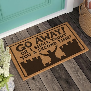 May include: A rectangular tan coir doormat featuring a dark brown border and a silhouette of a castle with a figure standing in front. The text reads: GO AWAY! OR I SHALL TAUNT YOU A SECOND TIME! in a bold, dark brown, sans-serif font.