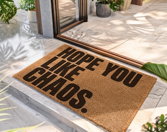 Puede incluir: Un felpudo rectangular de fibra de coco color canela con el texto negro HOPE YOU LIKE CHAOS. La alfombra tiene una textura fibrosa y áspera, colocada sobre un suelo de baldosas claras frente a una puerta, con plantas verdes cerca.
