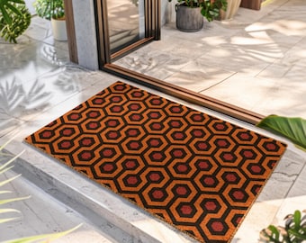 Puede incluir: Un felpudo rectangular con un patrón geométrico repetitivo de hexágonos entrelazados en tonos naranja, rojo oscuro y negro. El felpudo está colocado sobre un suelo de baldosas claras frente a una puerta de cristal con marco oscuro.