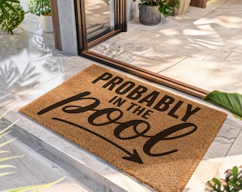Puede incluir: Un felpudo rectangular de fibra de coco color canela con el texto negro 'PROBABLY IN THE' en una fuente sans-serif negrita y la palabra 'pool' en una fuente cursiva grande y fluida, junto a una flecha negra que apunta hacia abajo.