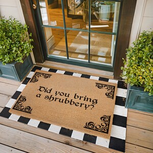 May include: A rectangular doormat with the text "Did you bring a shrubbery?" in black script. The mat is made of a natural fiber material and is bordered by a black and white striped pattern. Decorative corner accents add detail.