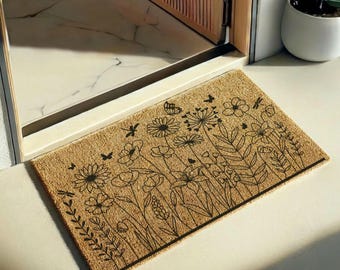 Puede incluir: Un felpudo rectangular de fibra de coco color canela con una ilustración de líneas negras de varias flores silvestres, hojas, mariposas y libélulas. El diseño cubre la superficie de la alfombrilla, situada sobre un suelo claro.