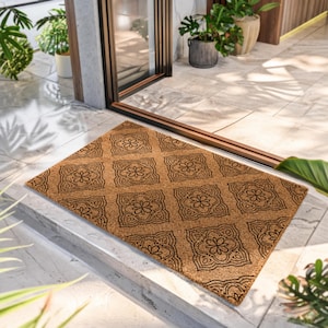 May include: A rectangular natural tan coir doormat featuring a repeating black geometric pattern of square tiles with central floral motifs. The mat has a coarse, fibrous texture and is positioned on a light-colored tiled floor surface.