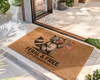 Puede incluir: Un felpudo rectangular de fibra de coco color canela con un mapache de dibujos animados que lleva gafas de sol rojas y azules y sostiene una bandera estadounidense. El texto dice FERAL & FREE 250th Independence, con fuegos artificiales y estrellas.