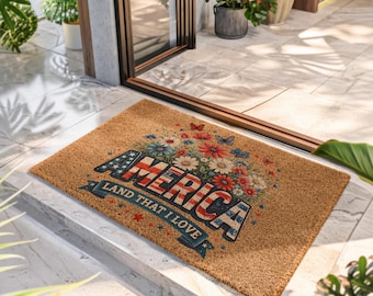 Puede incluir: Un felpudo rectangular de fibra de coco color canela con un arreglo floral de flores rojas, blancas y azules y mariposas sobre el texto AMERICA LAND THAT I LOVE en una fuente patriótica, con pequeñas estrellas rojas y azules en la superficie.