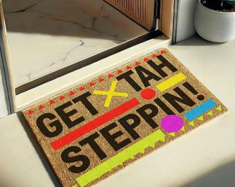 Puede incluir: Un felpudo rectangular de fibra de coco color canela con el texto negro GET TAH STEPPIN! decorado con formas geométricas en rojo, amarillo, azul y rosa. El diseño incluye pequeños triángulos rojos, una X amarilla, una línea roja y círculos.