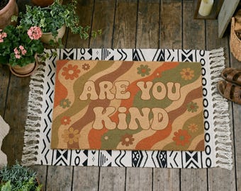 Puede incluir: Un felpudo rectangular con un patrón ondulado de inspiración retro en tonos naranja, marrón y verde. El texto ARE YOU KIND está impreso en letras grandes, negritas y de color claro en el centro, rodeado de pequeñas flores.