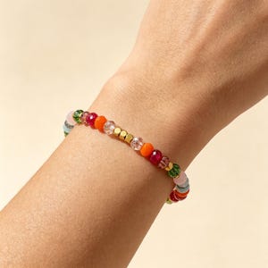 May include: A beaded bracelet featuring a sequence of multi-colored faceted beads in shades of orange, red, green, and light pink, interspersed with small metallic gold-toned cube spacers and clear crystal-like beads arranged in a repeating pattern.