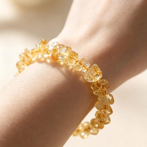May include: A stretch bracelet composed of numerous small, irregular, translucent yellow citrine gemstone chips. The stones are faceted and polished, creating a textured surface that reflects light, strung together on an elastic cord.