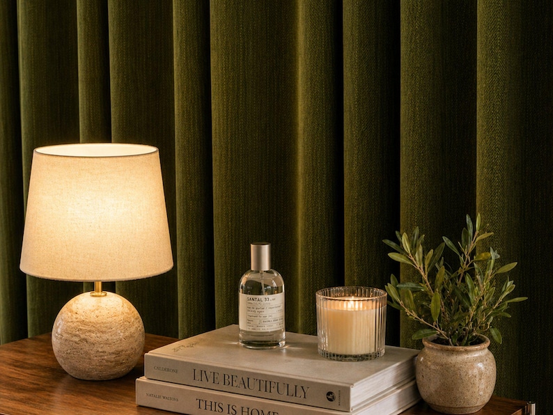 Peut inclure: Une lampe de table avec un abat-jour beige et une base sph&eacute;rique textur&eacute;e est pos&eacute;e &agrave; c&ocirc;t&eacute; d'un flacon de parfum, d'une bougie allum&eacute;e et d'une petite plante en pot. Les objets sont sur des livres avec les titres "LIVE BEAUTIFULLY" et "THIS IS HOME".