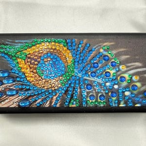 Puede incluir: Un estuche rectangular negro con una superficie superior decorativa adornada con un diseño de pluma de pavo real. El patrón está embellecido con numerosas cuentas pequeñas, coloridas y facetadas en tonos azul, verde y dorado, creando un efecto texturizado.